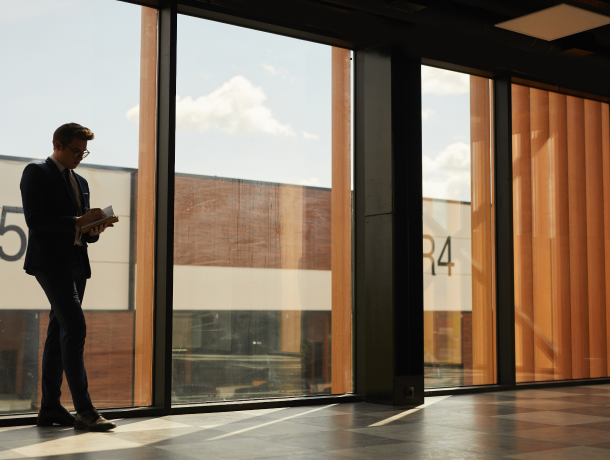 Man standing inside building next to floor to ceiling windows, reading a book